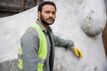 Poważny hinduski pracownik w kamizelce i rękawicy o wysokiej widoczności, stojący w pobliżu rzeźby betonowej na zewnątrz składowiska odpadów, koncepcja sortowania i recyklingu odpadów