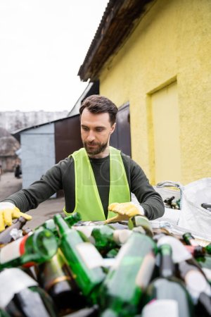 Bearded male worker in protective vest and gloves standing near blurred glass trash in outdoor waste disposal station, garbage sorting and recycling concept