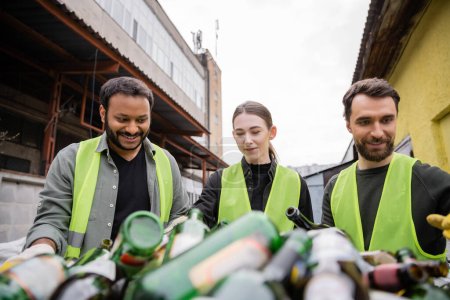 Positive interracial sorters in protective vests working with blurred glass trash in outdoor waste disposal station, garbage sorting and recycling concept