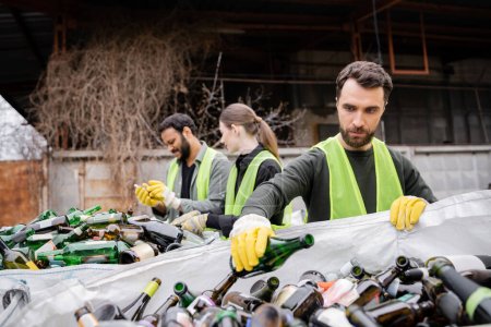 Brodatego pracownika w rękawiczkach ochronnych i kamizelce umieszczającego szklaną butelkę w worku w pobliżu rozmazanych współpracowników międzyrasowych w zewnętrznym składowisku odpadów, koncepcji sortowania i recyklingu odpadów