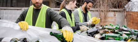 Bearded sorter in protective gloves and vest putting glass bottle in sack near blurred multiethnic colleagues in outdoor waste disposal station, garbage sorting and recycling concept, banner 