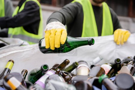 Cropped view of blurred worker in protective gloves and vest putting glass bottle in sack people in outdoor waste disposal station, garbage sorting and recycling concept