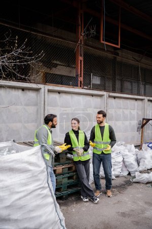Téléchargez les photos : Travailleur indien souriant en gilet haute visibilité et gants parlant à des collègues joyeux tout en se tenant près des sacs avec des ordures dans la station d'élimination des déchets extérieurs, le tri des ordures et le concept de recyclage - en image libre de droit