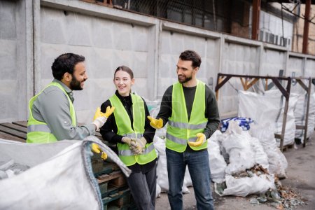 Trabajador positivo en chaleco de alta visibilidad y guantes hablando con colegas multiétnicos cerca de sacos con basura en la estación de eliminación de residuos al aire libre, clasificación de basura y concepto de reciclaje