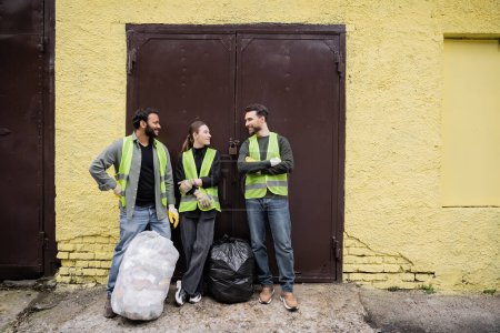 Sonrientes trabajadores multiétnicos en chalecos y guantes de protección hablando cerca de bolsas de basura y puerta de la estación de eliminación de residuos al aire libre, clasificación de basura y el concepto de reciclaje