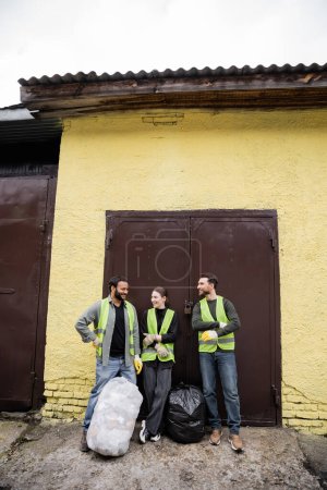 Positive multiethnische Arbeiter in Sicherheitswesten und Handschuhen im Gespräch, während sie in der Nähe von Müllsäcken und Tür der Müllentsorgungsstation im Freien stehen, Mülltrennung und Recyclingkonzept
