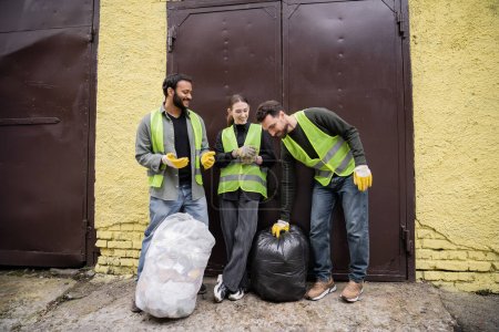 Gut gelaunte Arbeiter in Warnweste und Handschuhen stellen Müllsäcke in die Nähe internationaler Kollegen und der Tür der Müllentsorgungsstation im Freien, Mülltrennung und Recyclingkonzept