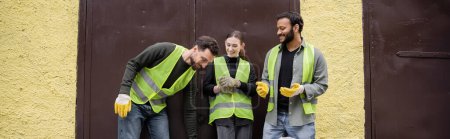 Positive multiethnische Arbeiter in Warnwesten und Handschuhen im Gespräch, während sie in der Nähe der Tür der Müllentsorgungsstation im Freien stehen, Müllsortier- und Recyclingkonzept, Transparent 