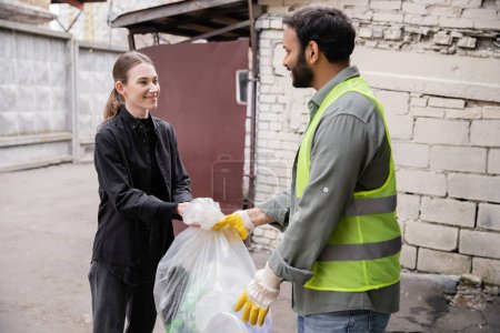 Smiling young volunteer giving plastic bag with trash to indian worker in protective vest and gloves outdoors in waste disposal station, garbage sorting and recycling concept