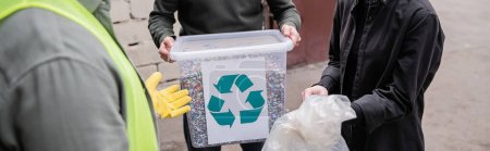 Ausgeschnittene Ansicht eines Arbeiters in Schutzweste und Handschuh, der in der Nähe von Freiwilligen steht, mit Müll in der Außenentsorgungsstation, Müllsortier- und Recyclingkonzept, Transparent 
