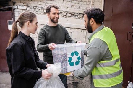 Bénévoles souriants donnant des ordures à un travailleur indien en gilet haute visibilité dans une station d'élimination des déchets de plein air, concept de tri et de recyclage des déchets
