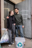 Cheerful volunteers hugging and holding trash bag near bin while standing next to door of waste disposal station outdoors, garbage sorting and recycling concept Sweatshirt #658271846