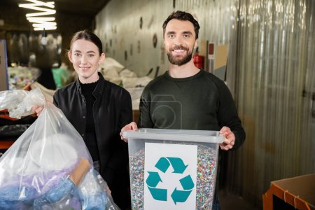 Cheerful volunteers looking at camera while holding trash bin and bag and standing together in blurred waste disposal station, garbage sorting and recycling concept