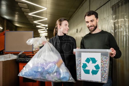 Smiling volunteer holding trash bag near man with bin and recycle sign in blurred waste disposal station at background, garbage sorting and recycling concept