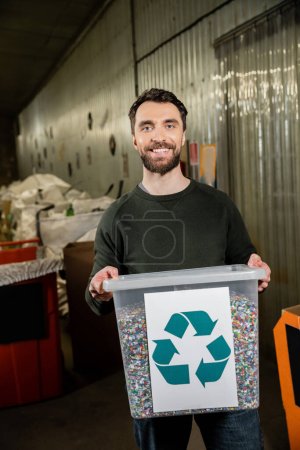 Voluntario alegre y barbudo mirando a la cámara mientras sostiene la papelera con el cartel de reciclaje en la estación de eliminación de residuos en el fondo, clasificación de basura y el concepto de reciclaje