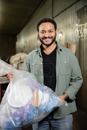 Uśmiechnięty indyjski wolontariusz patrzący w kamerę, trzymający plastikową torbę ze śmieciami w niewyraźnej stacji utylizacji odpadów w tle, koncepcję sortowania śmieci i recyklingu