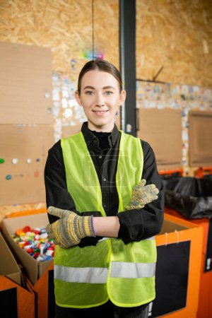 Smiling worker in high visibility vest and gloves looking at camera and crossing arms while standing near blurred plastic caps in box waste disposal station, garbage sorting and recycling concept