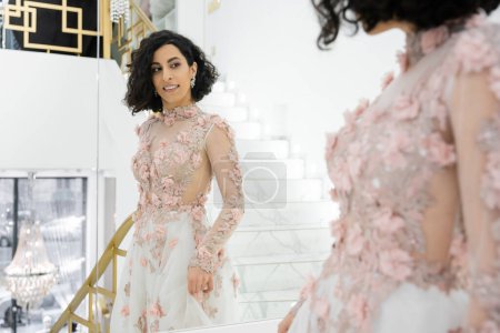 brunette femme du Moyen-Orient avec des cheveux ondulés debout dans une robe de mariée magnifique et florale tout en regardant miroir dans le salon de mariée luxueux, mariée heureuse, charmante et élégante, réflexion, shopping