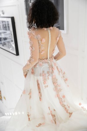 Rückansicht der brünetten Frau aus dem Nahen Osten mit welligem Haar, die in einem floralen Hochzeitskleid im Inneren eines luxuriösen Brautsalons wandelt, charmante und elegante Braut, verschwommene Fotografie an weißer Wand