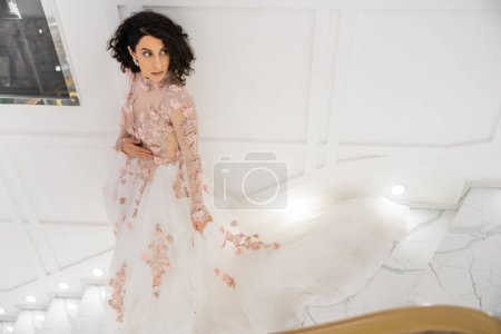 Hochwinkelaufnahme der brünetten Frau aus dem Nahen Osten mit welligem Haar, die in einem wunderschönen und floralen Hochzeitskleid mit Schleppe steht, während sie in einem luxuriösen Brautsalon wegschaut, Braut, weiße Wand 