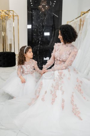 Photo for Happy and brunette middle eastern woman with wavy hair looking at cute girl and smiling near white wedding dresses in bridal salon, floral, mother and daughter, happiness, wedding day, shopping - Royalty Free Image
