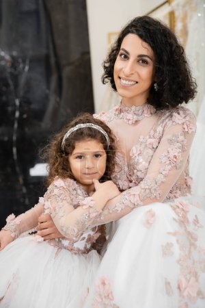 brunette middle eastern woman with wavy hair hugging cute girl and smiling near white wedding dresses in bridal salon, floral, mother and daughter, happiness, shopping, wedding day 