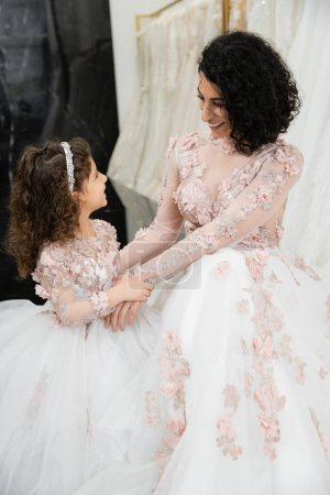 brunette middle eastern woman with wavy hair looking at girl and smiling near white wedding dresses in bridal salon, floral, mother and daughter, happiness, wedding day, shopping, bonding 