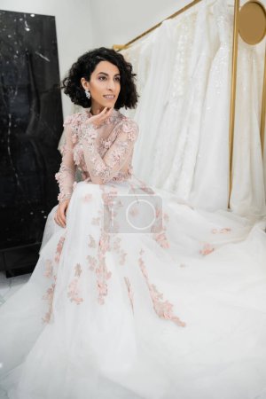 feliz mujer de Oriente Medio con el pelo ondulado sentado en vestido de novia hermosa y floral cerca borrosa y vestido blanco dentro de lujoso salón de novia, encantador y elegante, novia-a-ser
