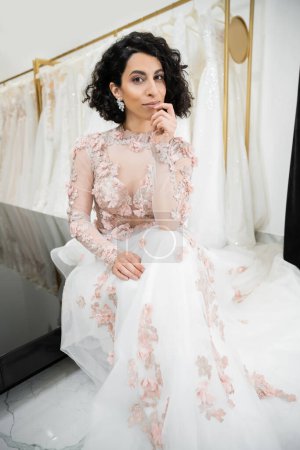 brünette Frau aus dem Nahen Osten mit welligem Haar, die in einem wunderschönen und floralen Hochzeitskleid in der Nähe eines verschwommenen und weißen Kleides in einem luxuriösen Brautsalon sitzt, einkaufen geht, die zukünftige Braut in die Kamera schaut 