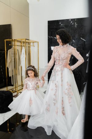 happy middle eastern woman with brunette wavy hair in stunning wedding dress holding hands with daughter in cute floral attire while standing in bridal salon, shopping, golden accents