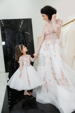 cheerful middle eastern woman with wavy hair in stunning wedding dress holding hands with daughter in cute floral attire while standing in bridal salon, shopping, luxurious, golden accents  Stickers 658421422