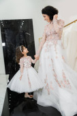 cheerful middle eastern woman with wavy hair in stunning wedding dress holding hands with daughter in cute floral attire while standing in bridal salon, shopping, luxurious, golden accents  t-shirt #658421422