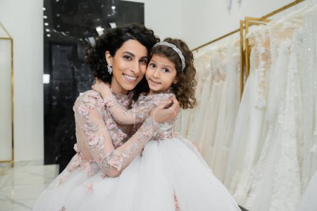 brunette middle eastern woman with wavy hair in stunning wedding dress hugging cute daughter in cute floral attire in bridal salon, shopping, happiness, special moment, togetherness 