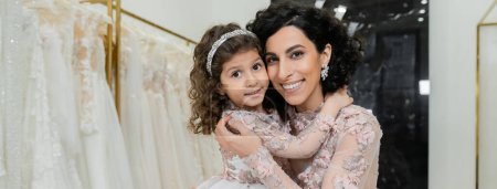 brunette middle eastern woman with wavy hair in stunning wedding dress hugging cute daughter in cute floral attire in bridal salon, shopping, happiness, special moment, togetherness, banner