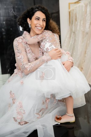 cheerful middle eastern bride with wavy hair in stunning wedding dress hugging girl in cute floral attire in bridal salon, shopping, happiness, special moment, togetherness, blurred white dresses