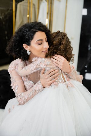 charming middle eastern bride with wavy hair in stunning wedding dress hugging daughter in cute floral attire in bridal salon, shopping, happiness, special moment, togetherness, blissful 