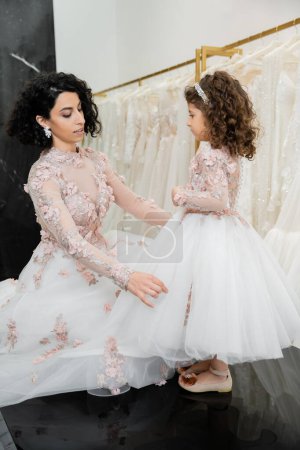 Photo for Charming middle eastern bride with brunette wavy hair in wedding dress adjusting tulle skirt of daughter in cute floral attire in bridal salon, shopping, special moment, togetherness - Royalty Free Image