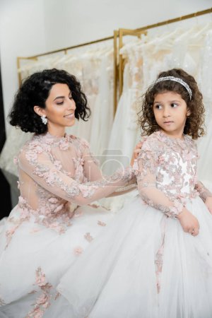 Photo for Charming middle eastern bride with brunette wavy hair in wedding dress adjusting cute floral dress of daughter in bridal salon, shopping, special moment, togetherness, blurred white gown - Royalty Free Image