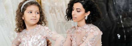 elegant middle eastern bride with brunette wavy hair in wedding dress adjusting cute floral attire of daughter in bridal salon, shopping, special moment, blurred white gown, banner