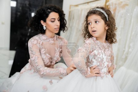 blurred middle eastern bride with brunette wavy hair in wedding dress adjusting cute floral dress of surprised daughter in bridal salon, shopping, special moment, hands on hips, white gown