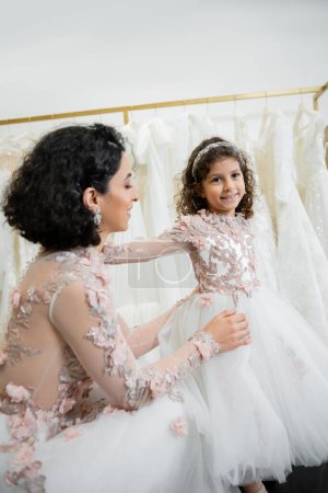 happy middle eastern woman with brunette wavy hair in floral wedding dress adjusting tulle skirt of daughter in cute attire in bridal salon, shopping, special moment, togetherness 