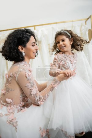 happy middle eastern bride with brunette wavy hair in floral wedding dress holding hands with smiling daughter in cute attire with tulle skirt in bridal salon, shopping, special moment 