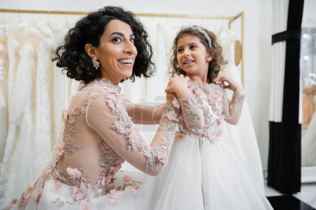 glückliche Frau aus dem Mittleren Osten mit brünetten Haaren im floralen Hochzeitskleid, lächelnde Tochter in niedlicher Kleidung mit Tüllrock im Brautsalon, Shopping, besonderer Moment, Zweisamkeit, Wegschauen 