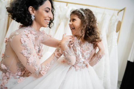 happy middle eastern woman with brunette hair in floral wedding dress hugging shoulders of smiling daughter in cute attire with tulle skirt in bridal salon, shopping, special moment, togetherness 