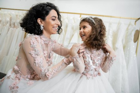 glückliche Frau aus dem Nahen Osten im floralen Hochzeitskleid umarmt Schultern lächelnder Mädchen in süßer Kleidung mit Tüllrock im Brautsalon, Shopping, besonderer Moment, Mutter und Tochter 