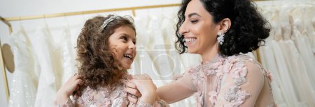 Photo for Happy middle eastern woman in floral wedding dress hugging shoulders of cheerful girl in cute attire in bridal salon, shopping, special moment, mother and daughter, bonding, banner - Royalty Free Image