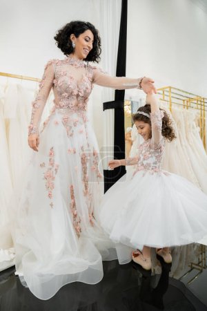 happy middle eastern woman in floral wedding dress dancing with smiling girl in cute attire with tulle skirt in bridal salon, shopping, special moment, mother and daughter, happiness 