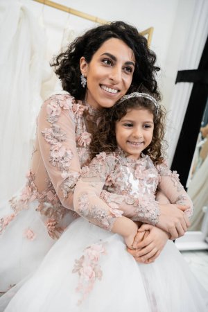 alegre mujer de Oriente Medio en vestido de novia floral abrazando chica feliz en traje lindo en salón de novia, compras, momento especial, madre e hija, felicidad 