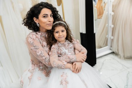 encantadora mujer de Oriente Medio en vestido de novia floral abrazando chica feliz en traje lindo en salón de novia, compras, momento especial, madre e hija, felicidad, mirando a la cámara