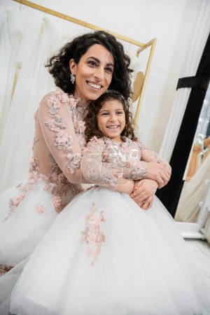 delightful middle eastern bride in wedding dress hugging happy girl in cute floral attire in bridal salon, shopping, special moment, mother and daughter, happiness, looking away
